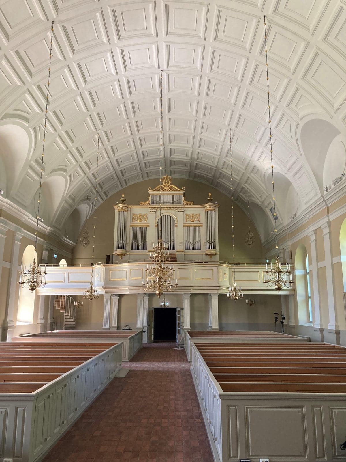 Kyrkorum i By kyrka med vit orgelläktare, förgylld orgel, ljuskronor och mittgång i tegel